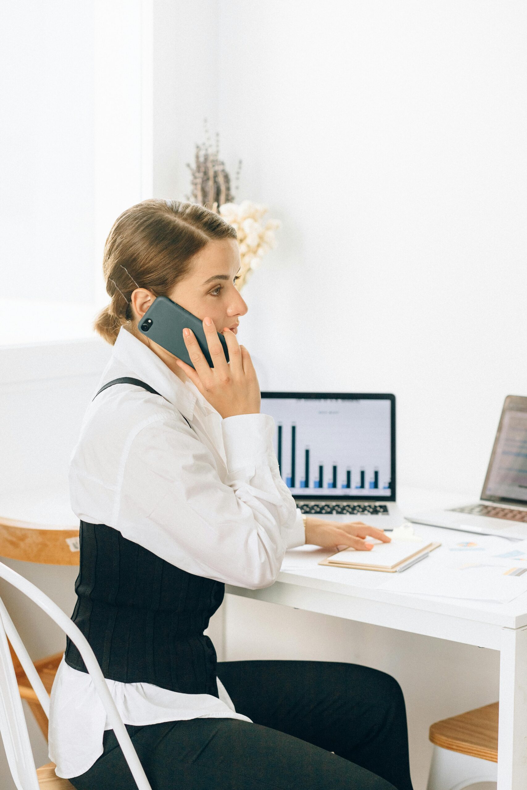 A businesswoman on the phone at her desk, working with a laptop displaying a bar chart and surrounded by documents.