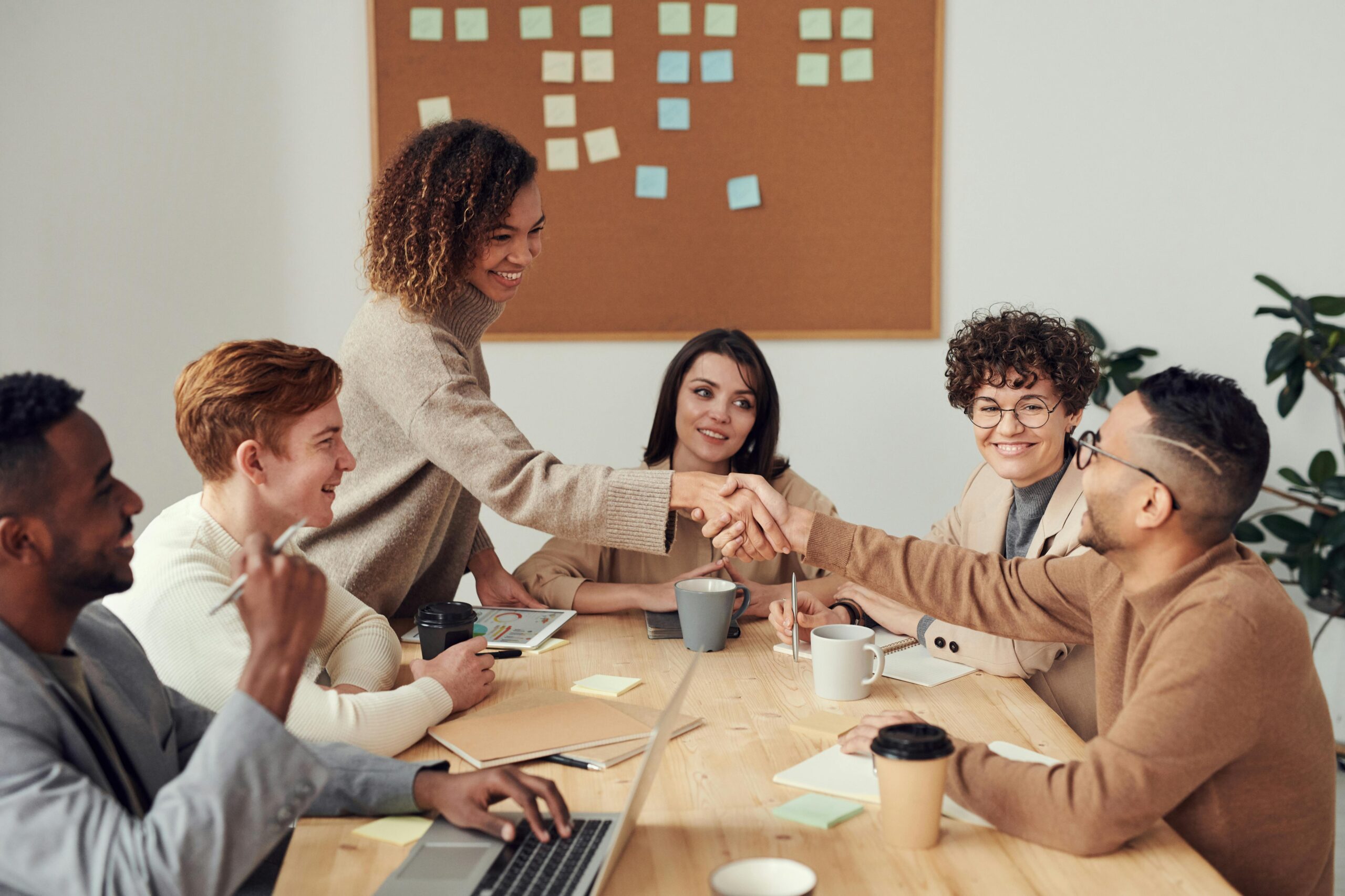 A diverse group of professionals in a meeting, shaking hands and discussing ideas with sticky notes on a board in the background.
