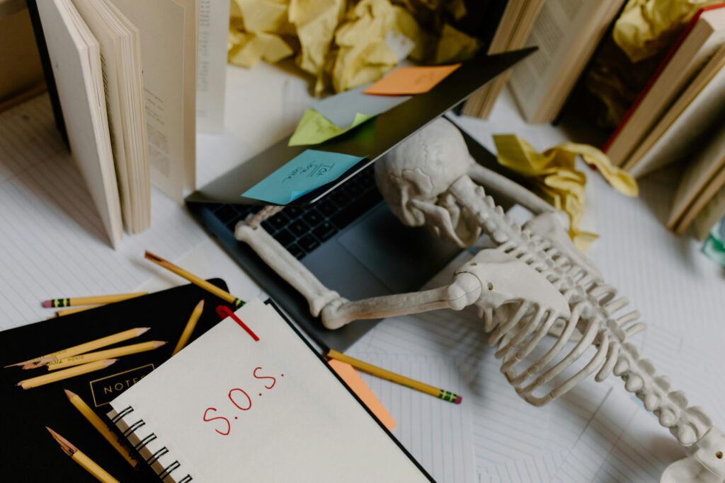 Skeleton slumped over laptop with S.O.S. note, symbolizing being overwhelmed by out-of-office reply chaos.