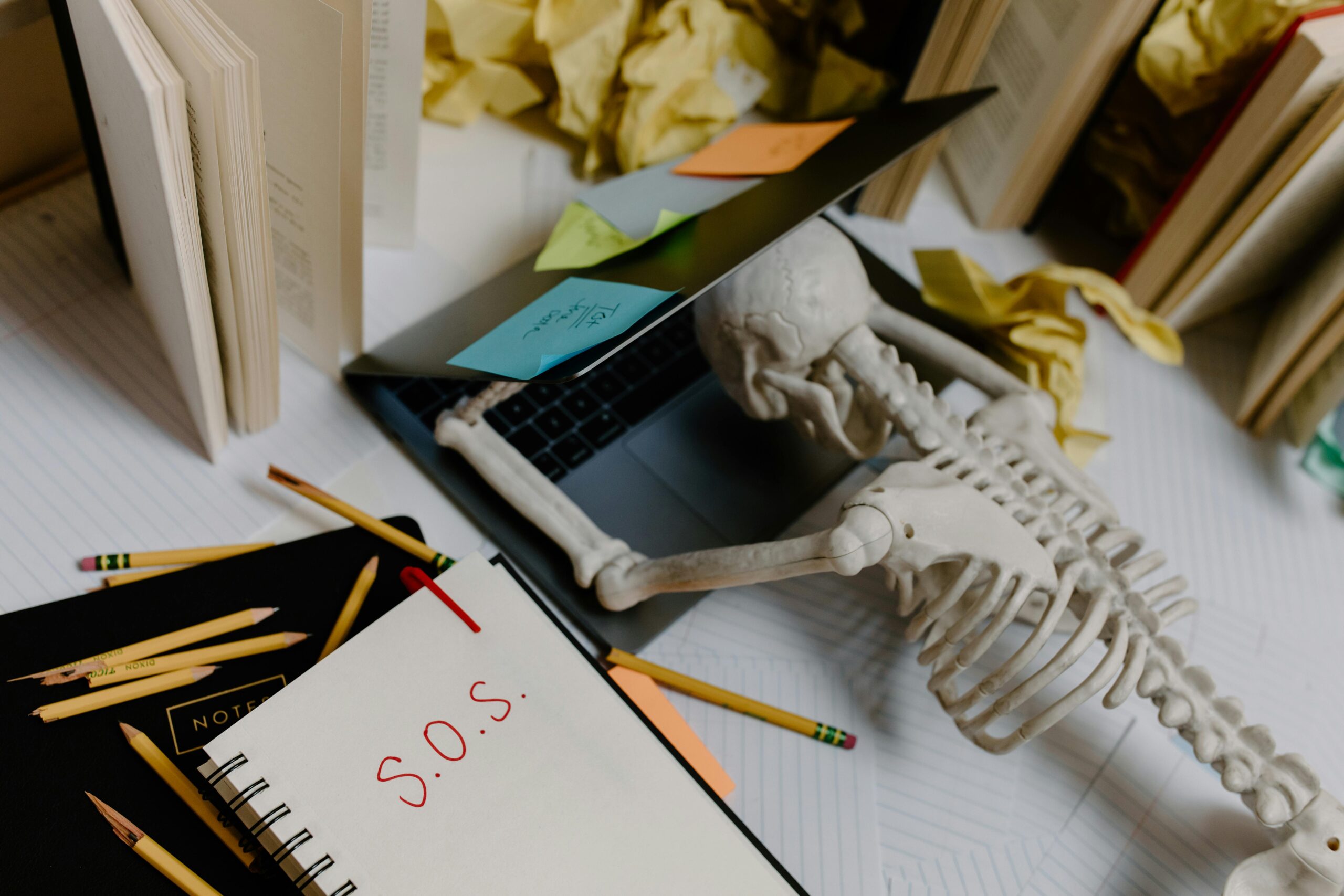 Skeleton slumped over laptop with S.O.S. note, symbolizing being overwhelmed by out-of-office reply chaos.