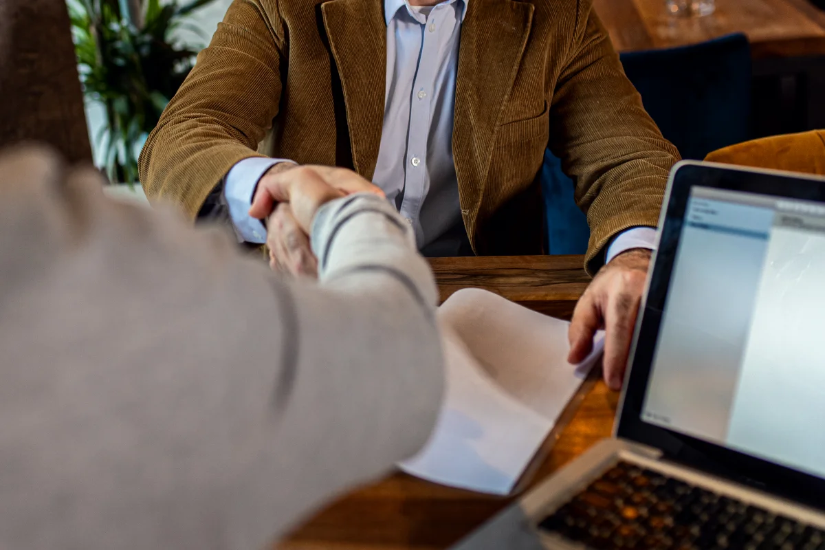 two businessmen in suits shaking hands with documents and laptop on table